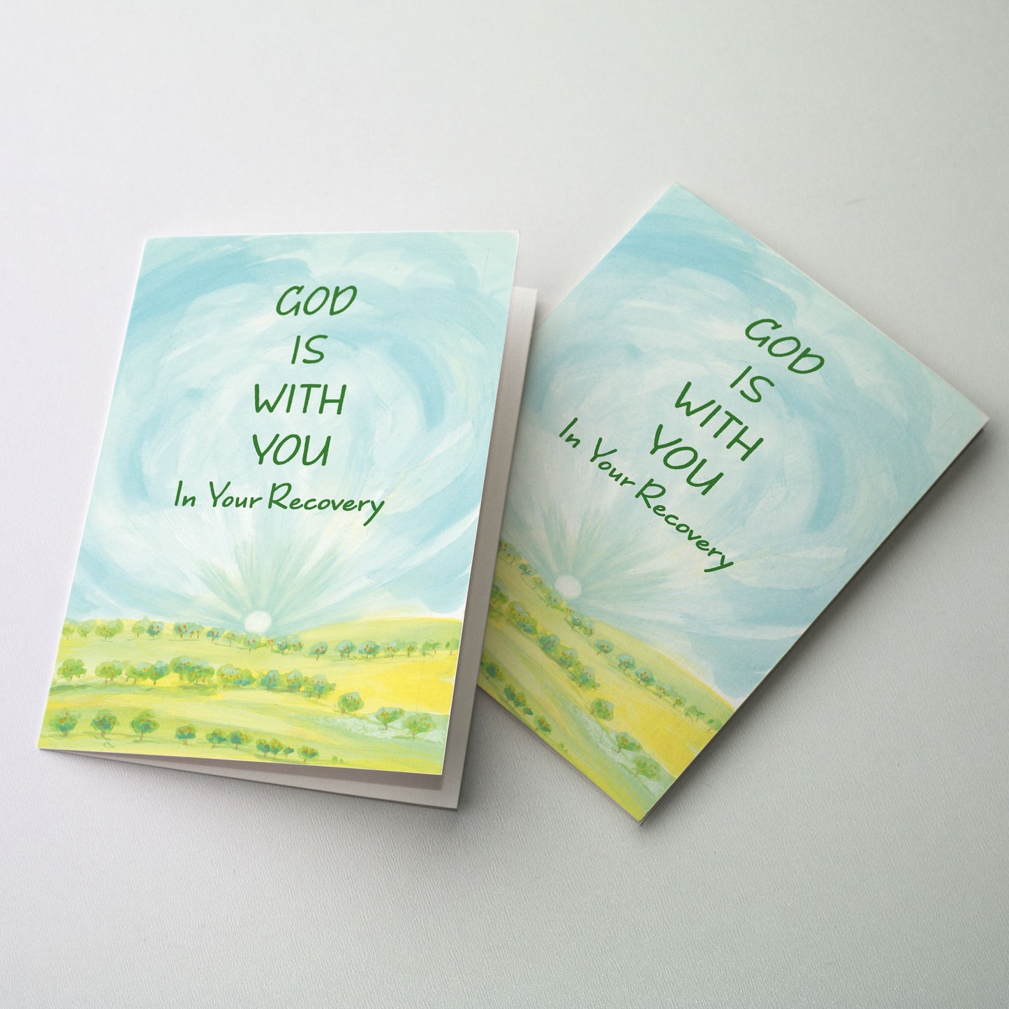 Watercolor landscape showing rows of fruit trees in the foreground with a rising sun on the horizon and active cloud formations in the blue sky above. The cards in “One Day at a Time” are reminders that God is there each step of the way during recovery, whether it’s rehab through a 12-step program or recovery from knee or hip surgery.