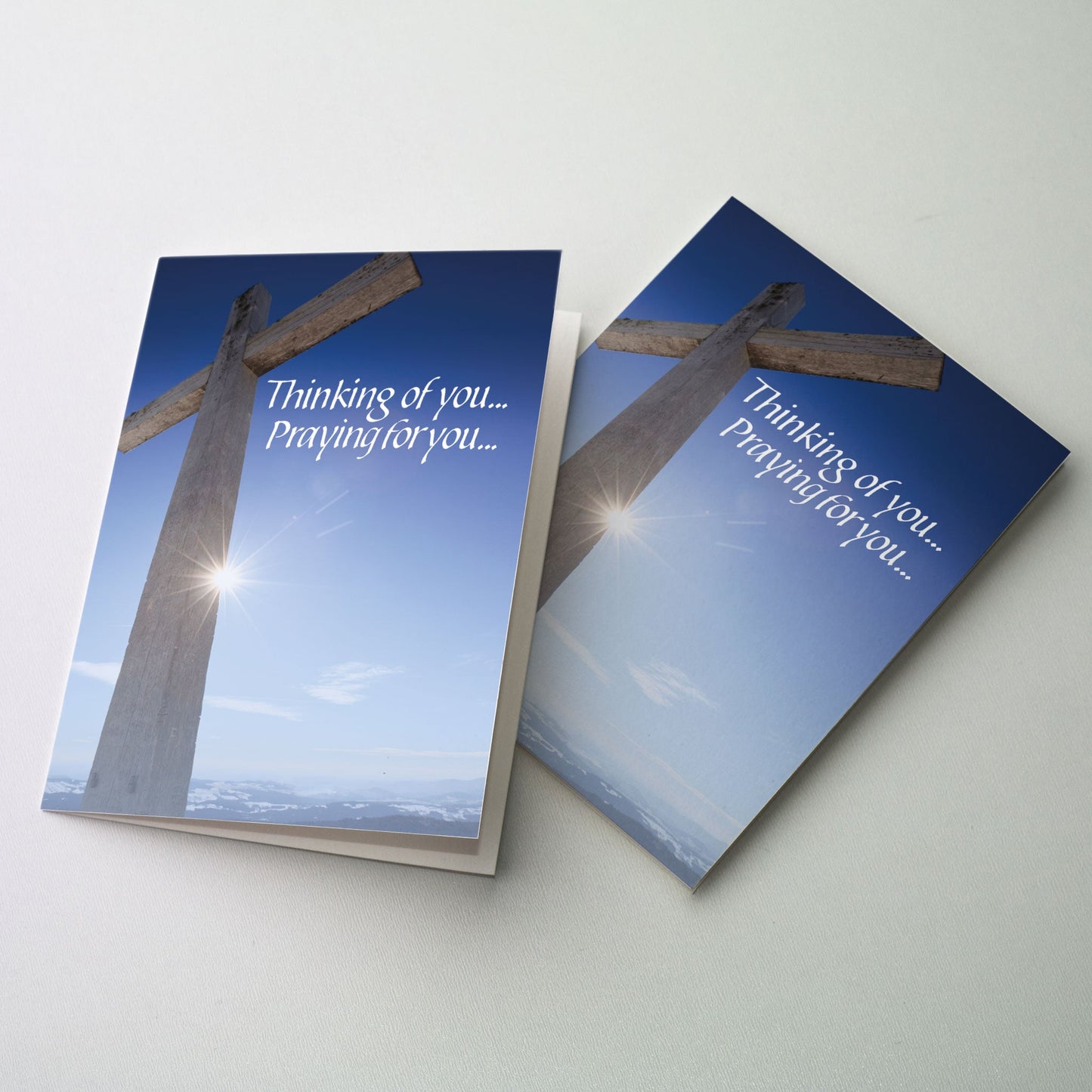 Photo of a rugged wooden cross atop a mountain against a brilliant blue sky with the sun visible behind the cross. Mountains are visible in the background.